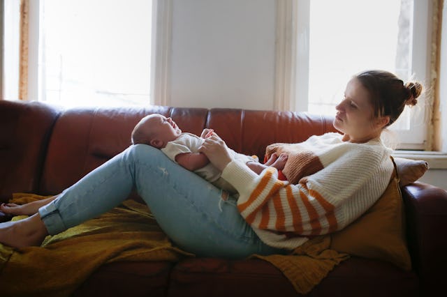 A mother sits comfortably on a couch, holding her baby. Natural light streams in, creating a warm, c...