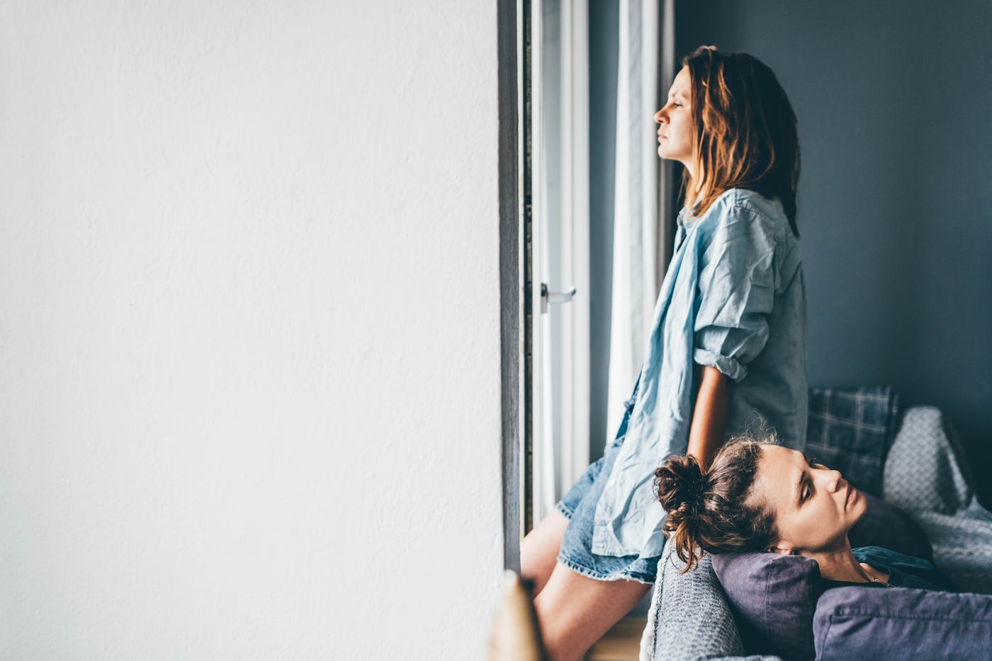 Two women are indoors; one stands by a window looking pensively out, while the other rests on a couch, gazing thoughtfully. Soft lighting enhances the mood.