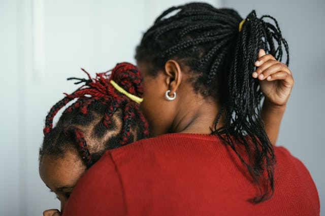 A person with braided hair embraces a child with red-streaked braids, both sharing a tender moment a...
