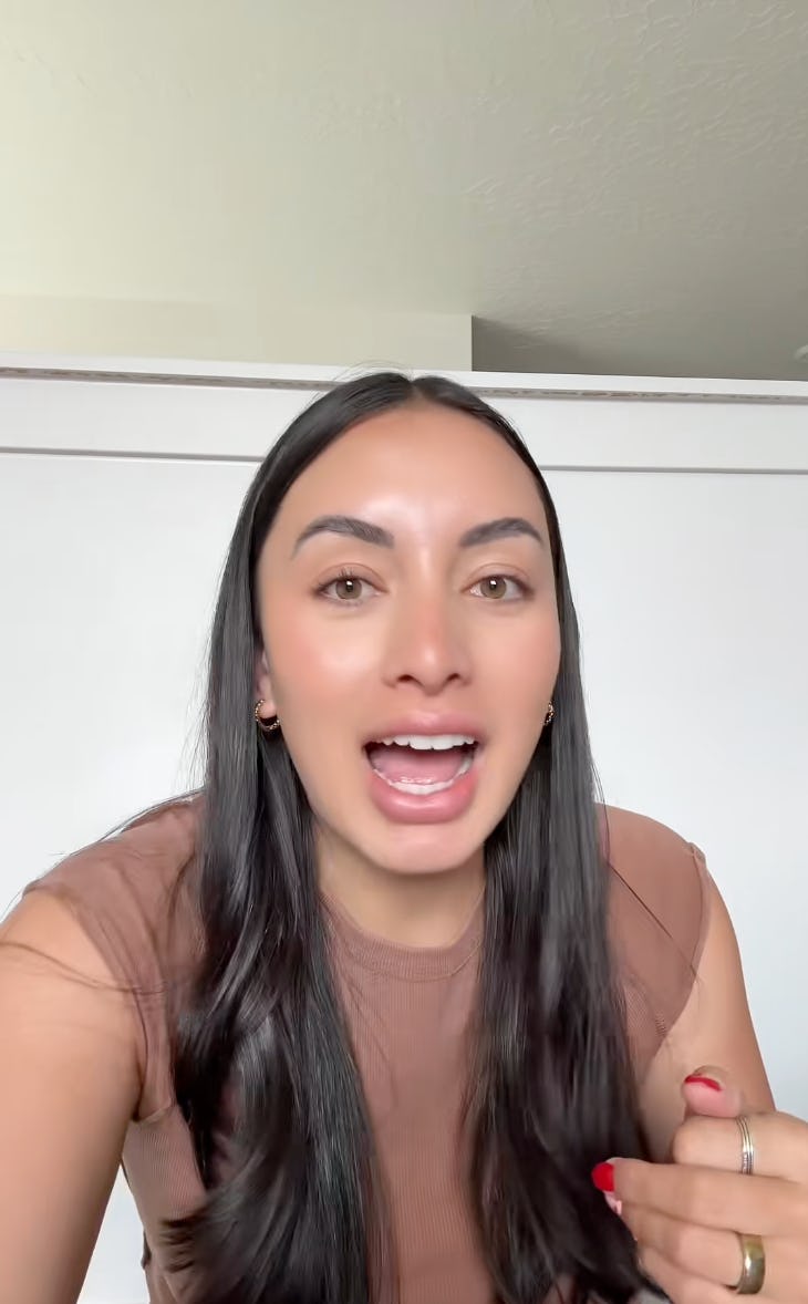 A smiling woman with long, dark hair, wearing a brown top, gestures while speaking. A white wall serves as the background, conveying a casual setting.