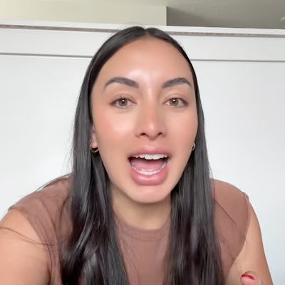 A smiling woman with long, dark hair, wearing a brown top, gestures while speaking. A white wall serves as the background, conveying a casual setting.