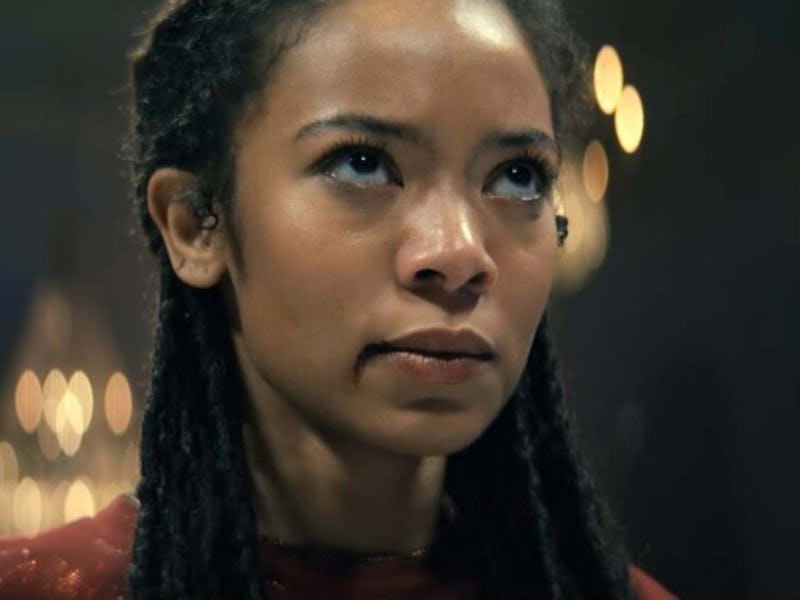 A close-up of a woman with dreadlocks, wearing a red outfit. She has an intense expression, with blu...