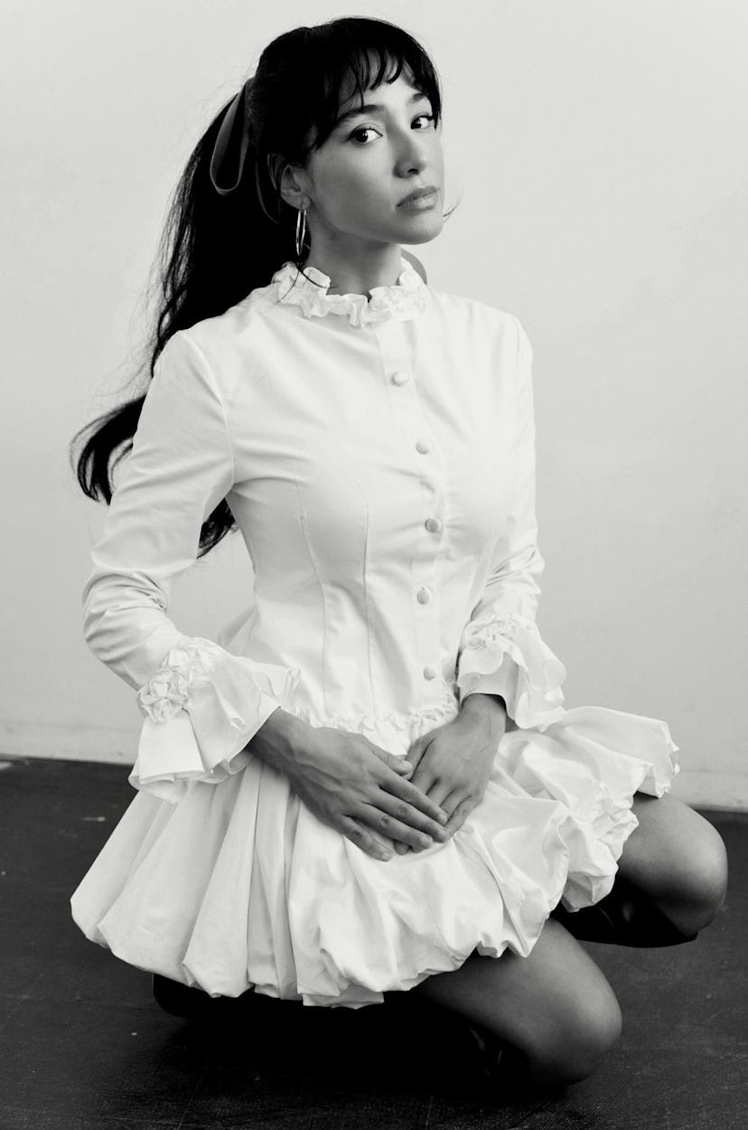 A young woman kneels in a white dress with ruffled sleeves and a high collar, her hair styled with l...