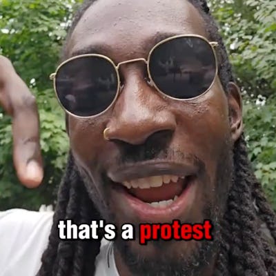 A smiling person with long hair and sunglasses gestures toward a vibrant green backdrop, speaking about a protest with enthusiasm.