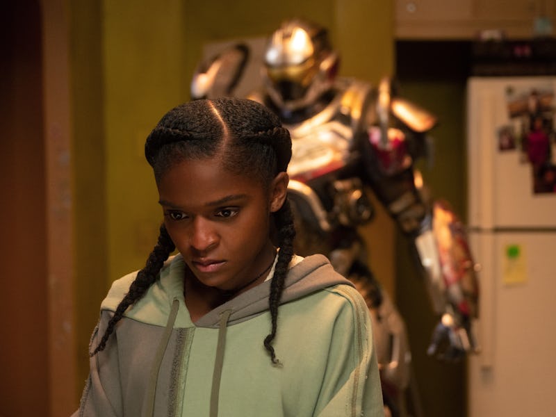 A girl with braided hair looks thoughtfully downward in a kitchen, while a large armored figure stan...