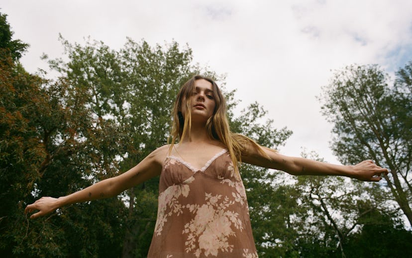 A woman stands in a natural setting, arms outstretched, wearing a sheer floral dress. Lush green tre...