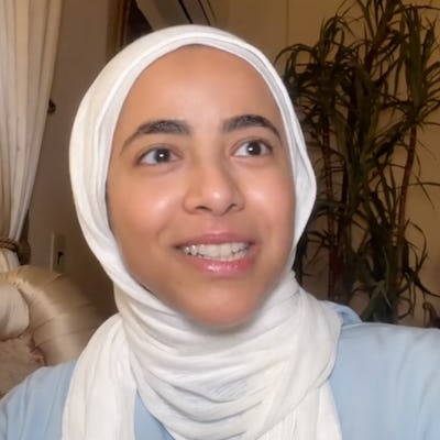 A young woman wearing a hijab smiles while speaking. She sits in a cozy indoor setting with plants and soft lighting in the background.