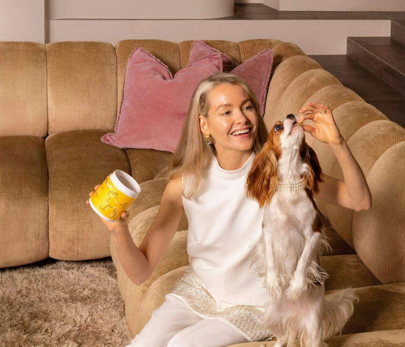 A smiling woman in a stylish outfit sits on a cozy couch, holding a yellow container and playfully feeding a small dog beside her.