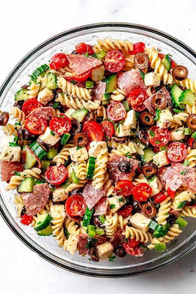 A colorful pasta salad in a clear bowl, featuring rotini, cherry tomatoes, olives, cucumbers, salami...