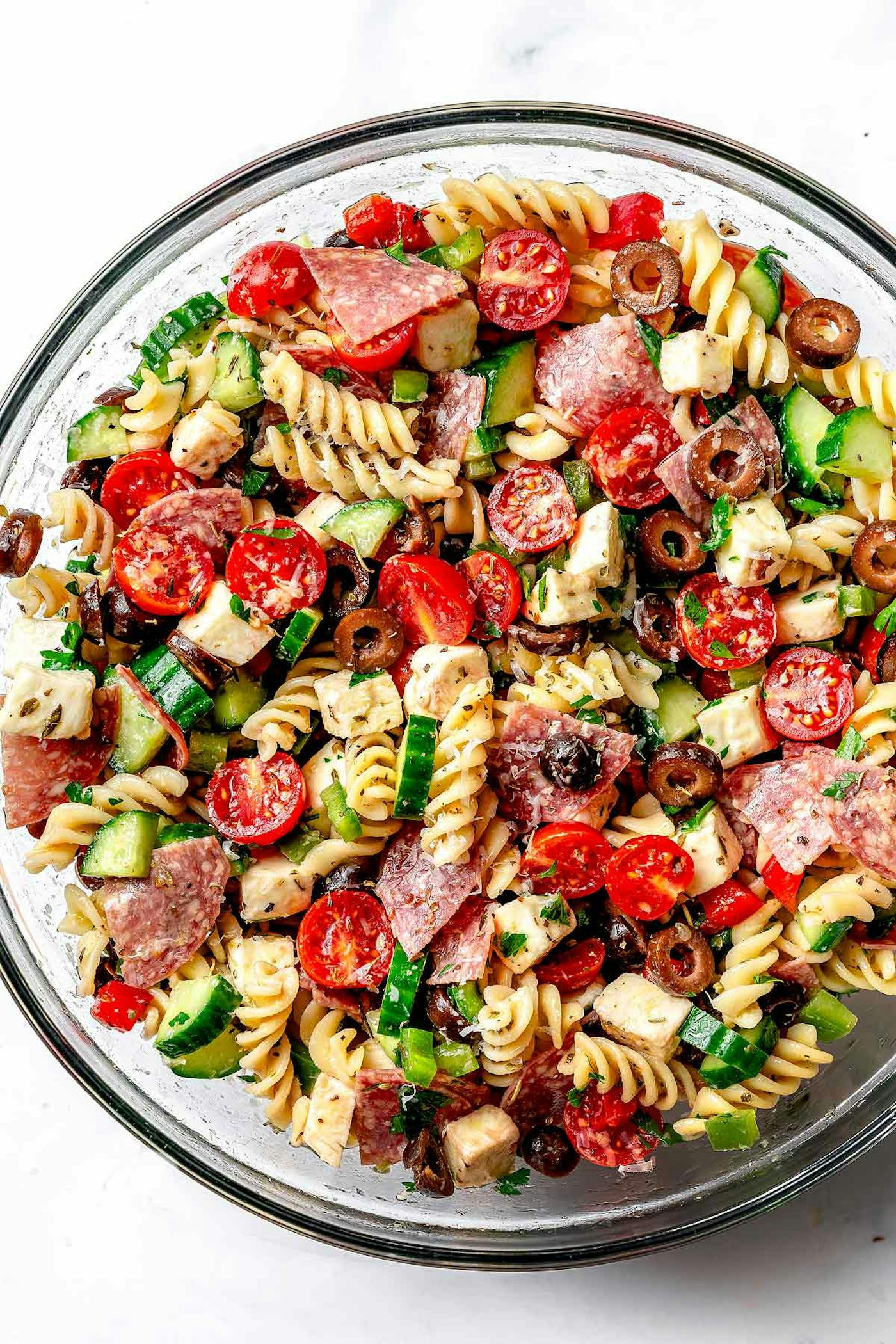 A colorful pasta salad in a clear bowl, featuring rotini, cherry tomatoes, olives, cucumbers, salami, and diced cheese, garnished with herbs.