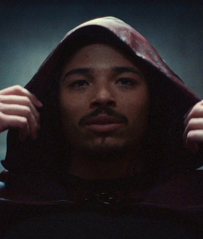 A young man wearing a dark hooded cloak looks thoughtfully at the camera, with his hands adjusting the hood, set against a moody background.
