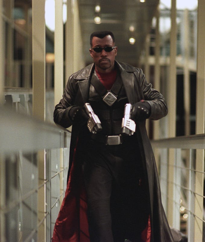 A stylish figure in a long black coat and red accents walks confidently down a modern corridor, wielding two silver guns. The dramatic lighting enhances the scene.