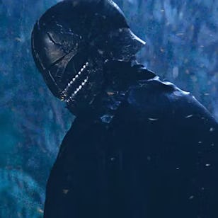A cloaked figure stands in a misty, darkened forest, wearing a menacing helmet with metallic features, surrounded by a mystical atmosphere.