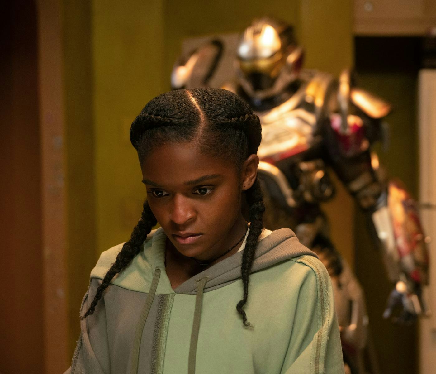 A young woman with braided hair looks pensively downward, wearing a light green hoodie. A large, armored figure stands behind her, partially obscured.