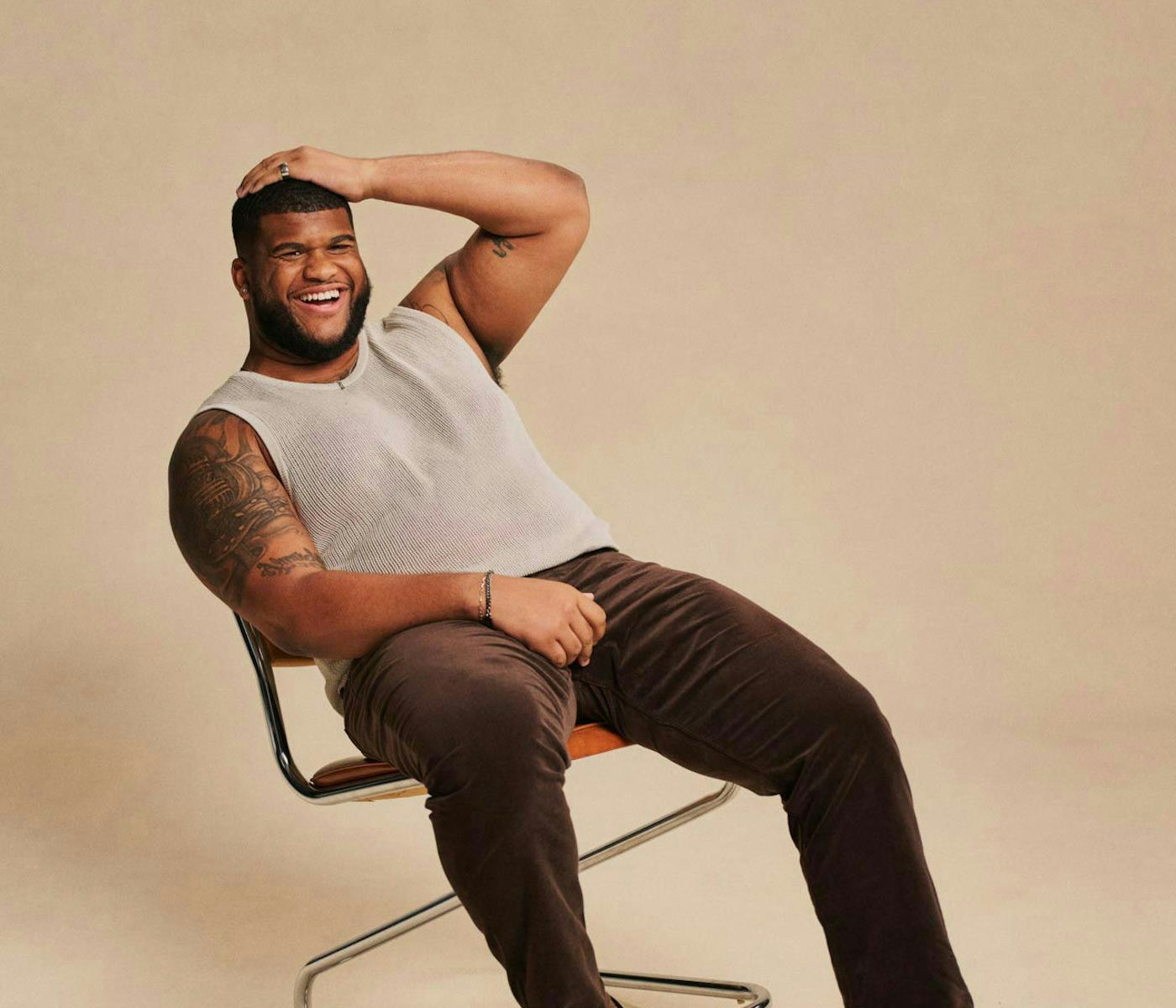 A smiling man with a tattoo sits comfortably on a chair, wearing a light tank top and brown pants, against a neutral background.