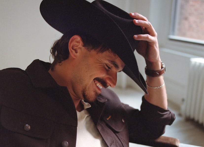 A man sits casually in a stylish black cowboy hat and a brown jacket, smiling warmly as he brushes h...
