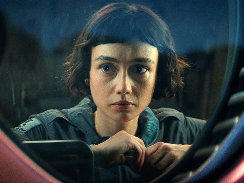 A close-up of a young woman with short hair, gazing intently through a round window. She appears con...