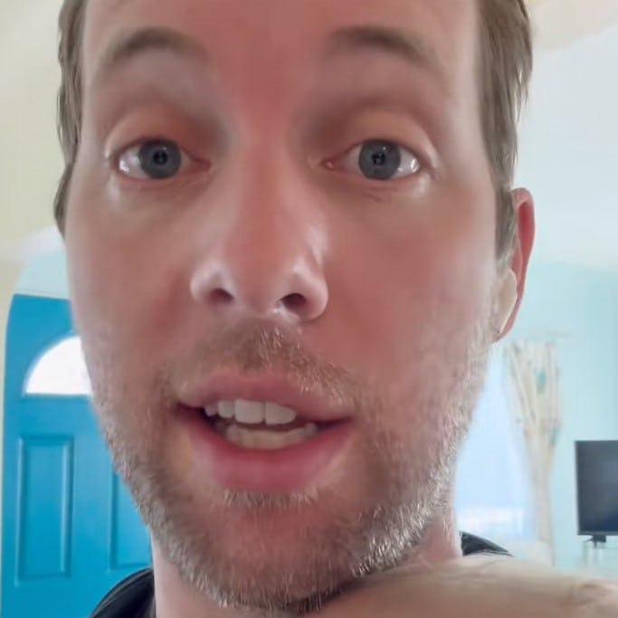 A man with light brown hair and blue eyes speaks directly to the camera, holding a baby in his arms, with a bright room in the background.