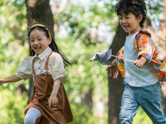 Two children joyfully run through a sunny park, holding colorful pinwheels. The background features trees and a relaxed outdoor atmosphere.