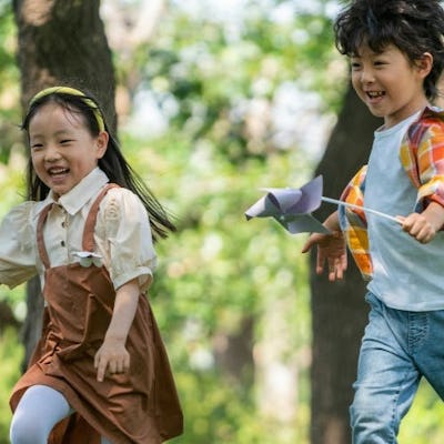 Two children joyfully run through a sunny park, holding colorful pinwheels. The background features trees and a relaxed outdoor atmosphere.