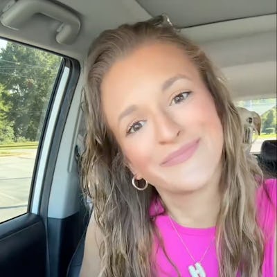 A young woman with long, wavy hair sits in a car, smiling at the camera. She wears a bright pink top, a necklace, and casual shorts.
