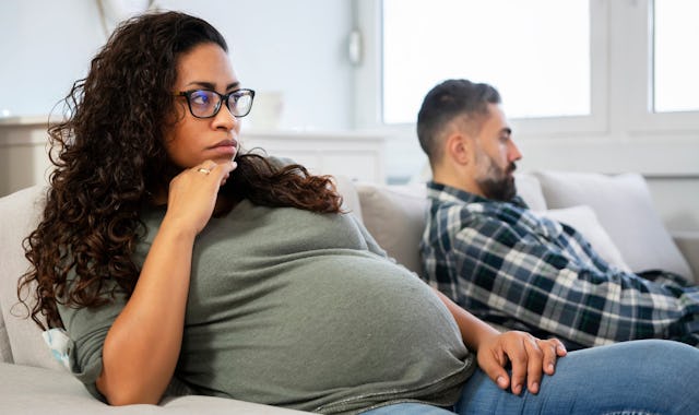 A pregnant woman with curly hair and glasses sits thoughtfully on a couch, while a man in a plaid sh...