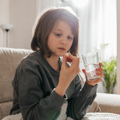 A young child sits on a couch, holding a clear cup while examining a small object in their hand, with sunlight streaming in from a window.