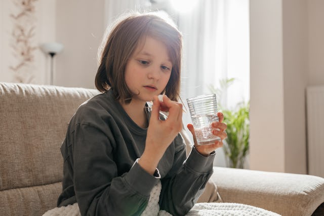 A young child sits on a couch, holding a clear cup while examining a small object in their hand, wit...