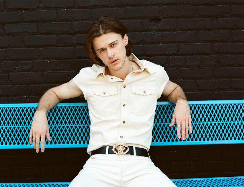 A young man in a cream-colored shirt and matching pants sits casually on a blue metal bench against ...