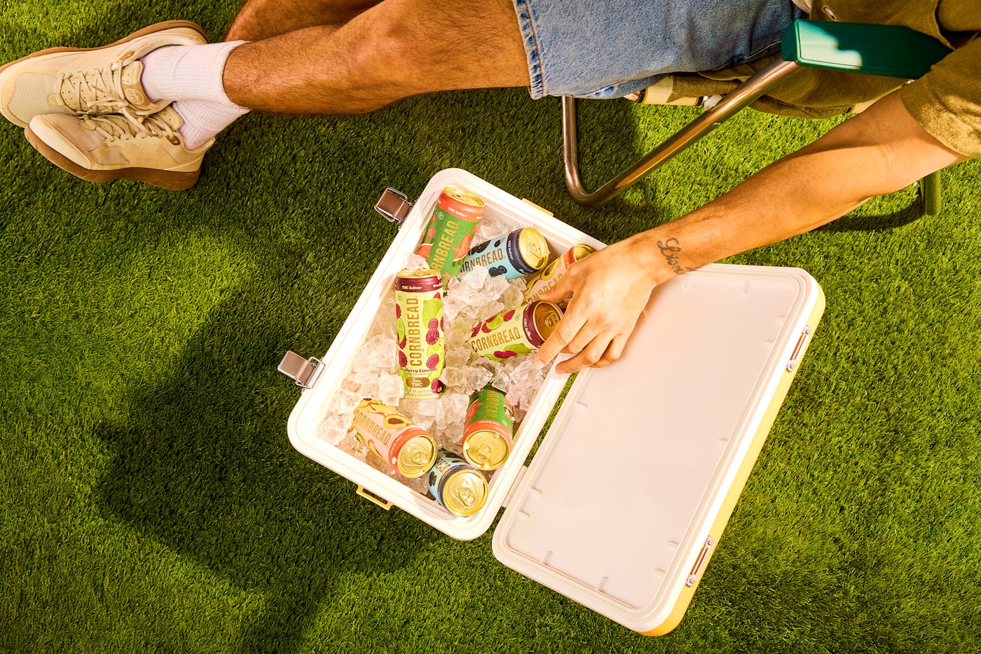 A person sits on green grass beside a cooler filled with ice and assorted colorful drinks, with their hand reaching into the cooler.