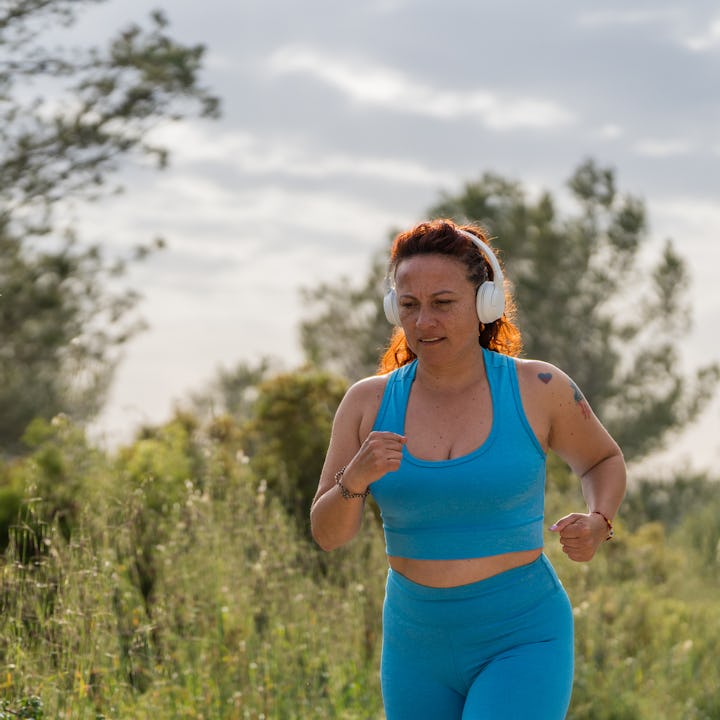 A woman in a blue workout outfit jogs through a lush green landscape, wearing headphones and enjoyin...