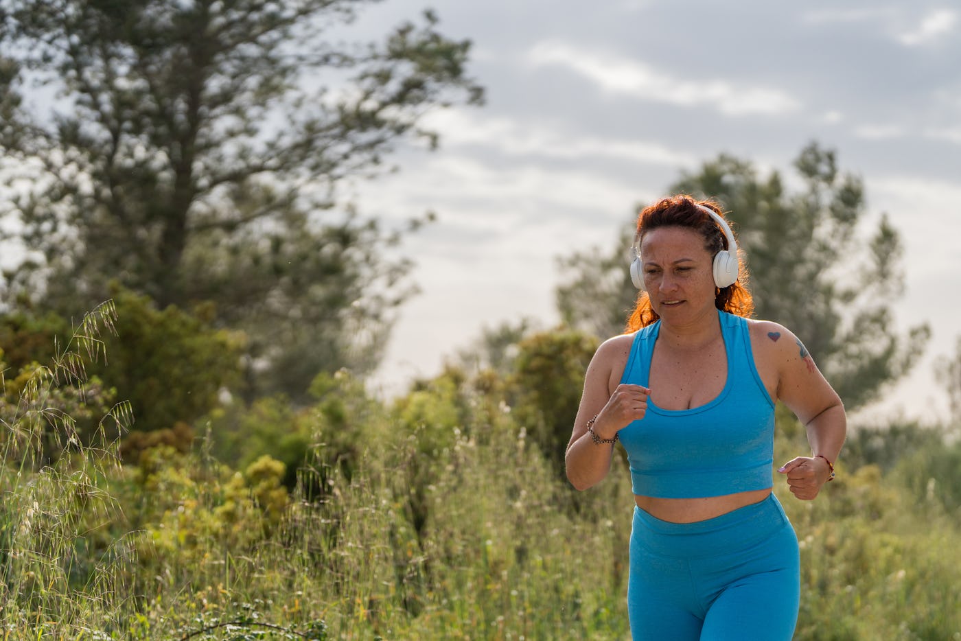 A woman in a blue workout outfit jogs through a lush green landscape, wearing headphones and enjoying the outdoors on a sunny day.