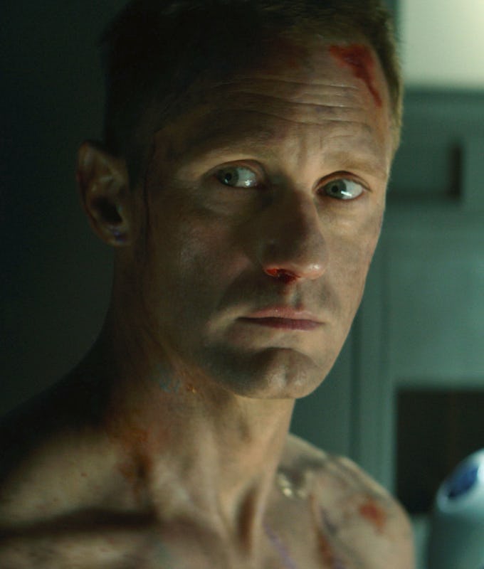 A close-up of a man with a distressed expression, featuring a cut on his nose and bruises on his bare torso, set against a dimly lit background.