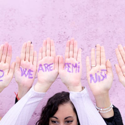A group of hands raised against a pink wall, displaying purple-painted words "WE ARE FEMI" and feminist symbols, promoting a message of feminism.