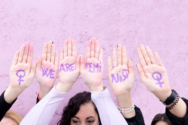 A group of hands raised against a pink wall, displaying purple-painted words "WE ARE FEMI" and femin...