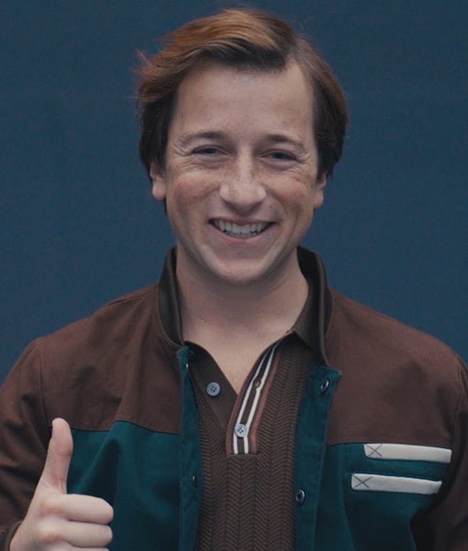 A young man with brown hair smiles and gives a thumbs-up, wearing a brown and teal jacket over a striped shirt, against a blue backdrop.