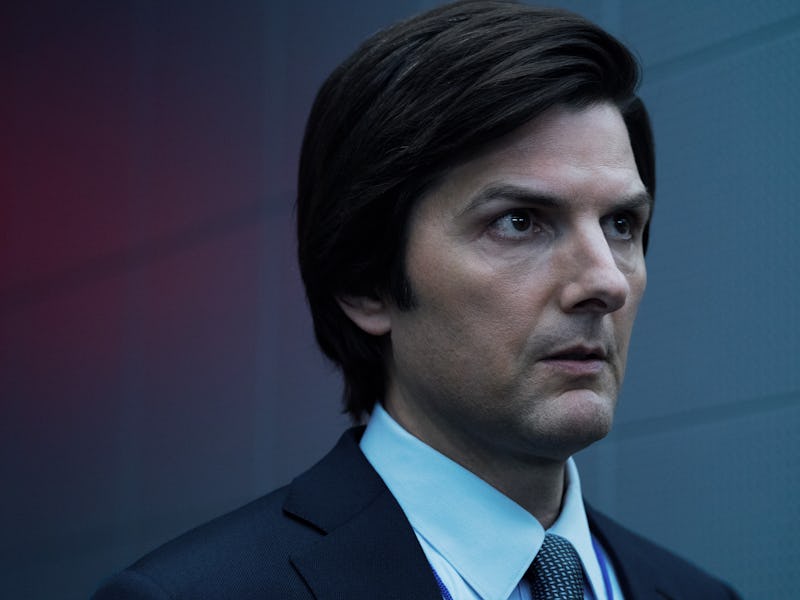 A man with dark hair and a serious expression wears a black suit and light blue shirt, standing agai...