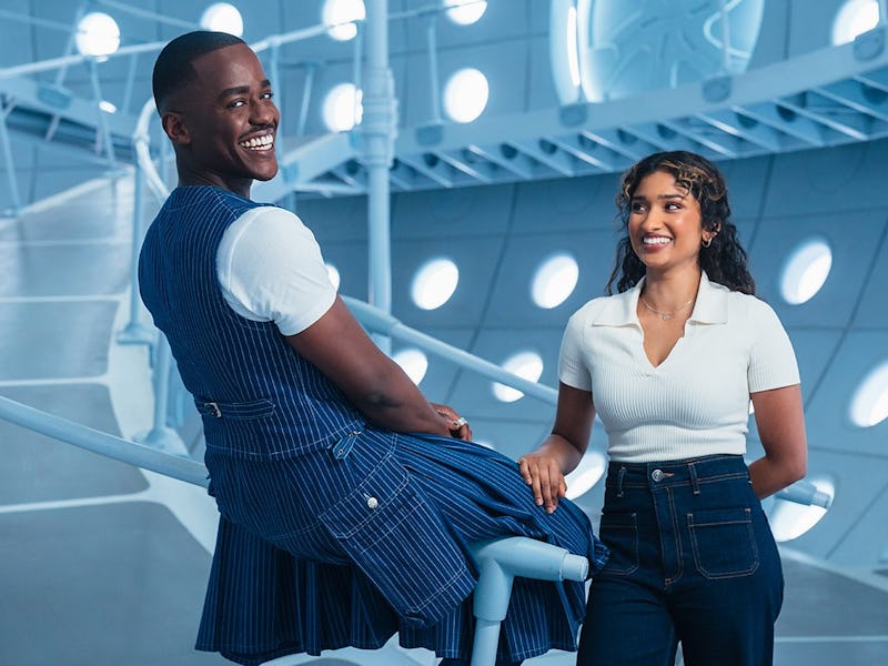 Two smiling individuals are posing in a modern, futuristic setting. One is seated, wearing a blue ou...