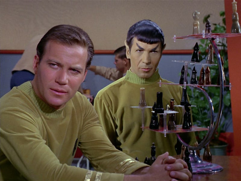 Two characters in matching gold uniforms are seated at a table. One leans back thoughtfully, while t...