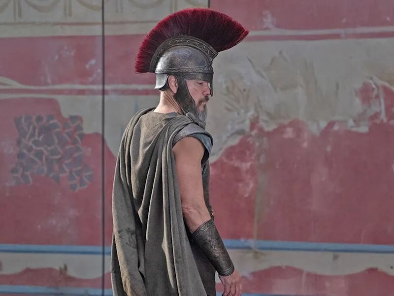 A warrior in ancient armor and a plumed helmet walks away, set against a faded mural with intricate ...