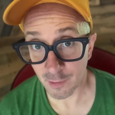 Steve Burns, wearing glasses, a yellow cap, and green shirt, gazes at the camera.