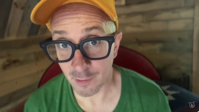 Steve Burns, wearing glasses, a yellow cap, and green shirt, gazes at the camera.