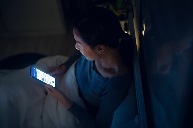 A woman sits in bed, illuminated by her phone's screen, wrapped in a blanket while browsing content ...