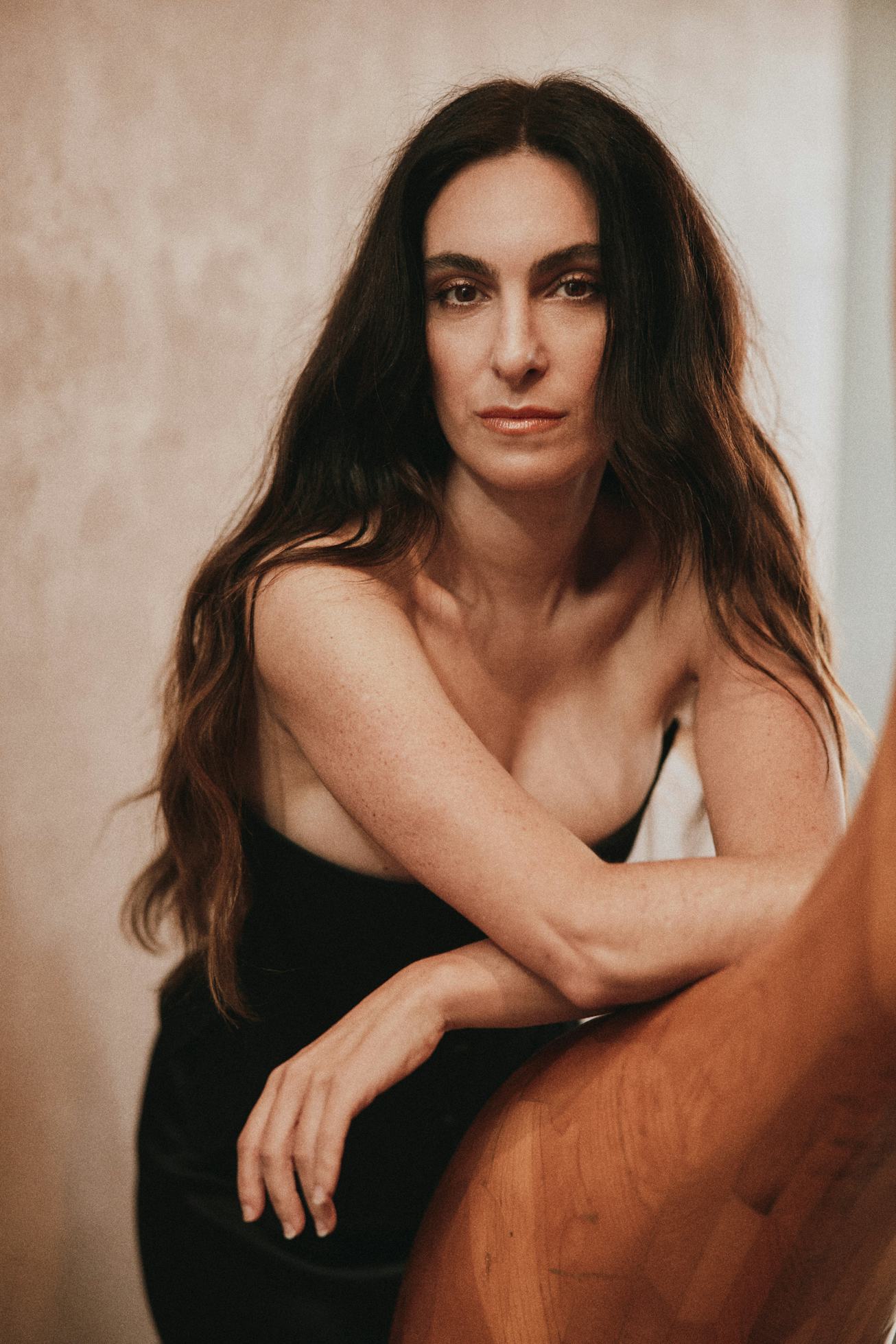 A woman with long, dark hair poses against a textured background, wearing a black dress. She leans o...