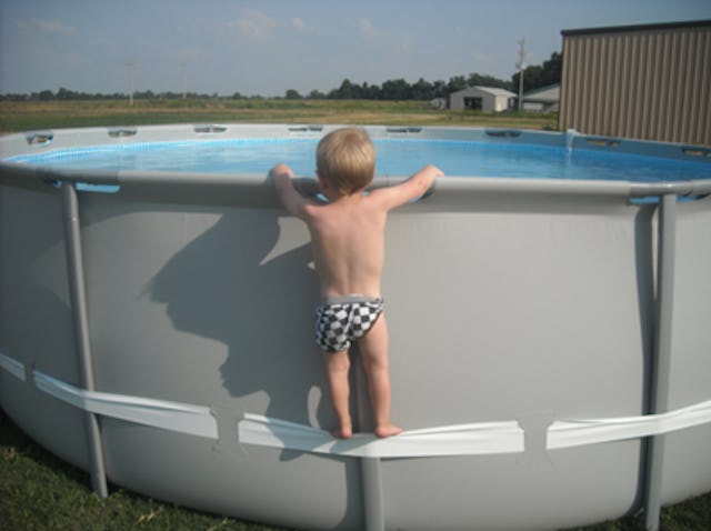 A toddler in a checkered swimsuit is climbing on the edge of a round above-ground pool, looking into...
