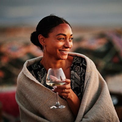 A woman wrapped in a blanket smiles while holding a cocktail glass, enjoying a relaxed moment outdoors, with a beach setting in the background.
