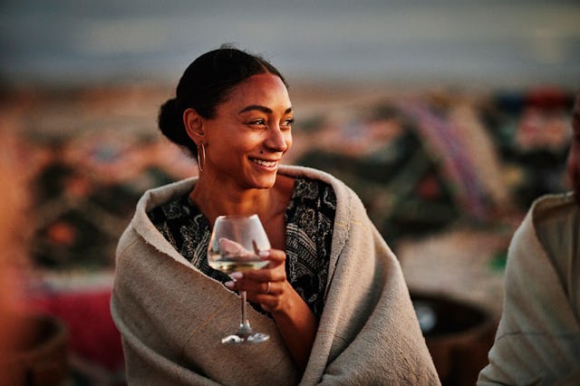 A woman wrapped in a blanket smiles while holding a cocktail glass, enjoying a relaxed moment outdoo...