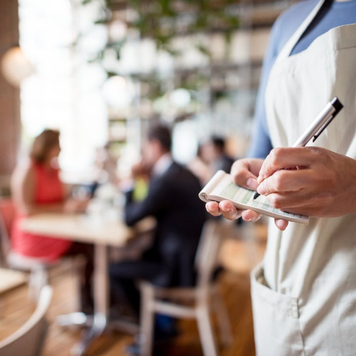 A waiter in a light apron takes notes on a notepad while standing in a bustling restaurant with cust...