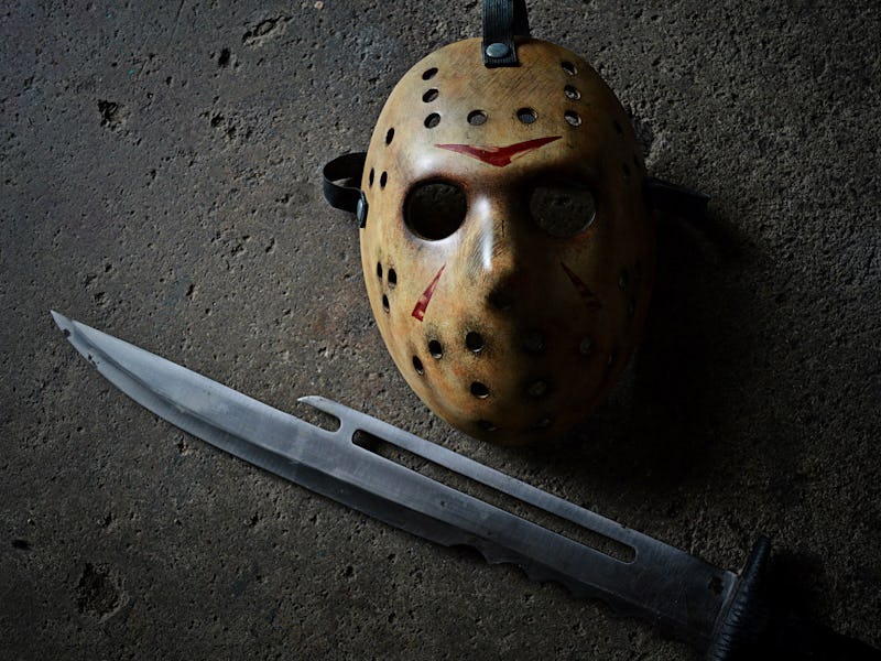 A worn, brown hockey mask with red markings lies on a rough surface, next to a long, curved knife, e...