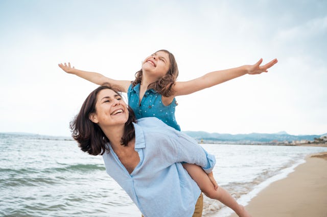 A joyful woman carries a smiling girl on her back at the beach, with arms outstretched, enjoying a c...
